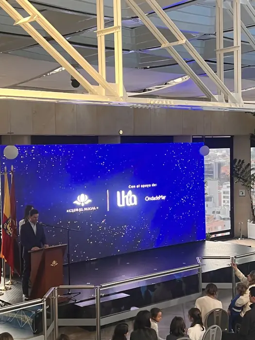 Pantalla LED azul de gran formato en un salón con ventanales, atril y banderas institucionales